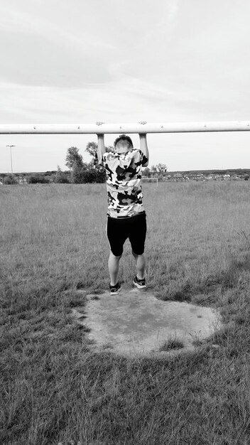 Full length of boy hanging from metallic rod on field against sky