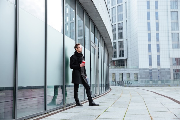 Full length of Businessman in warm clothes holding coffee in hand in the street and looking away.