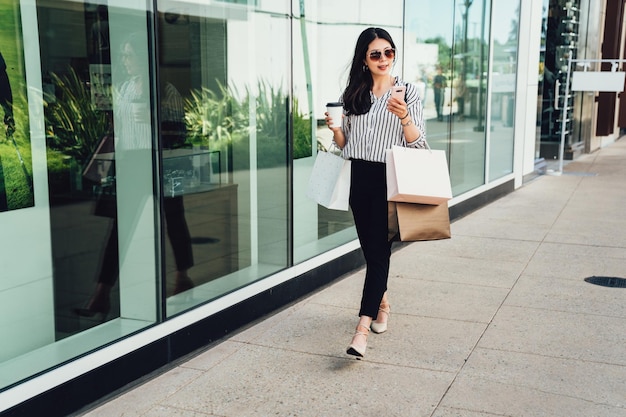 full length businesswoman holding coffee shopping for business carrying bags using cellphone replying email. young office lady walking in sunglasses on street outside leaving department store mall.