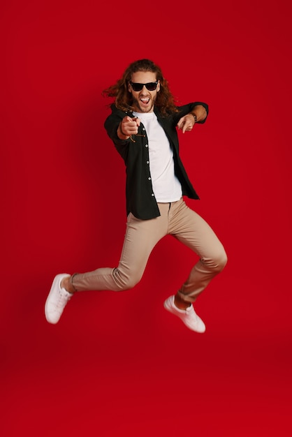 Full length of carefree young man in casual clothing smiling and pointing you while hovering against red wall