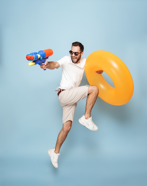 Full length of a cheerful excited man wearing blank t-shirt jumping isolated over blue wall, having fun with inflatable ring and water gun