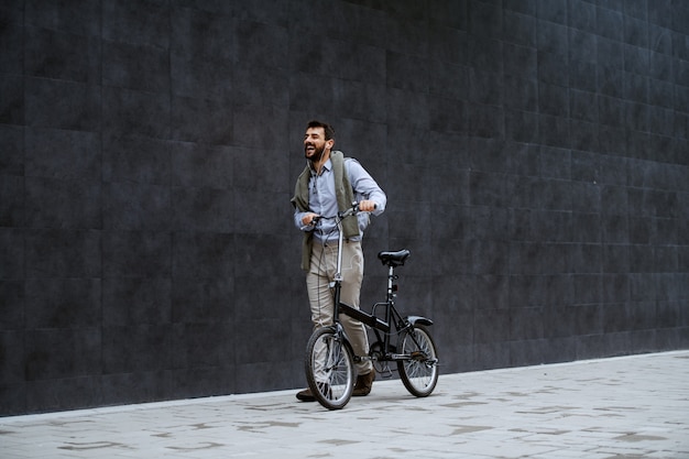 Full length of cheerful handsome caucasian fashionable man listening music and pushing bicycle. In background is gray wall.