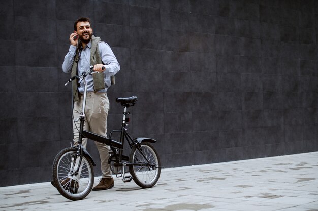 Full length of cheerful handsome caucasian fashionable man listening music and pushing bicycle. In background is gray wall.