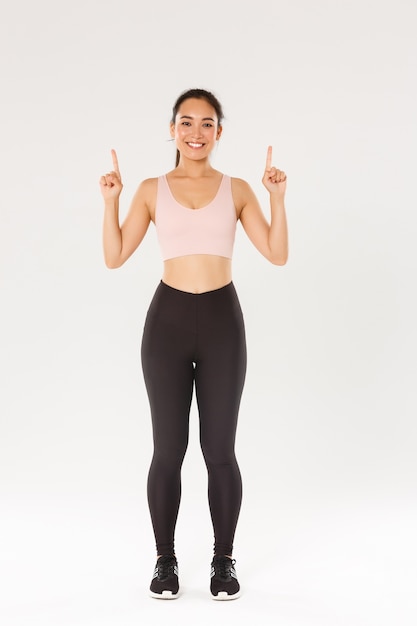 Full length of cheerful smiling and cute fitness girl, pointing fingers up, showing workout equipment advertisement.