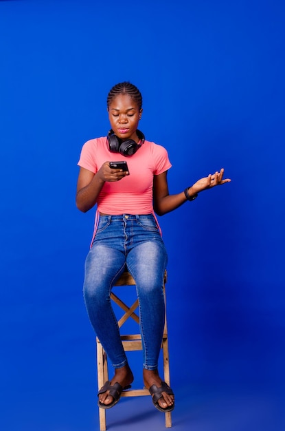 Full length of cheerful young black woman with headphones and cellphone listening to music or audio book sitting on chair