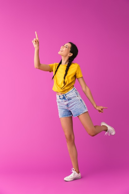 Per tutta la lunghezza di una giovane ragazza bruna allegra in piedi isolata sul rosa, puntando il dito verso l'alto