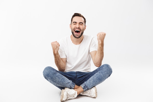 Full length of a cheerful young man sitting with legs crossed isolated over white wall, celebrating success