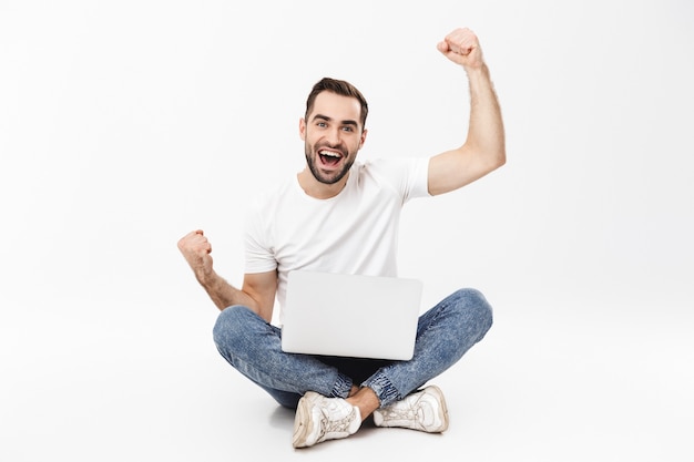 Full length of a cheerful young man sitting with legs crossed isolated over white wall, using mobile phone, celebrating