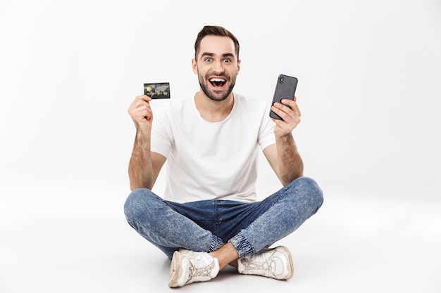 Full length of a cheerful young man sitting with legs crossed isolated over white wall, using mobile phone, showing credit card