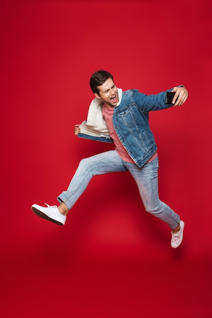 Full length of a cheerful young man wearing warm denim jacket jumping isolated over red wall, taking a selfie