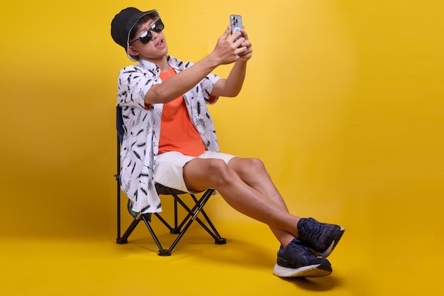 Full length cheerful young traveler tourist Asian guy deck chair doing selfie shot or recording vide
