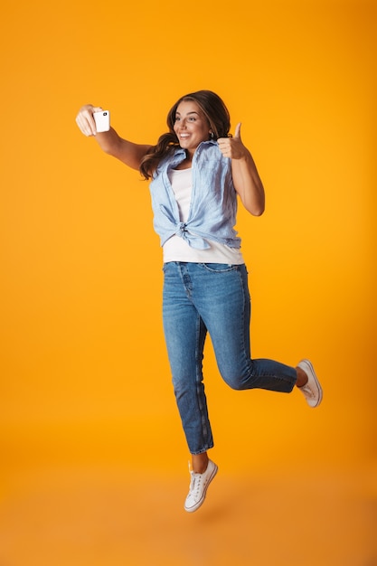 Full length of a cheerful young woman jumping isolated, taking a selfie with outstretched hand