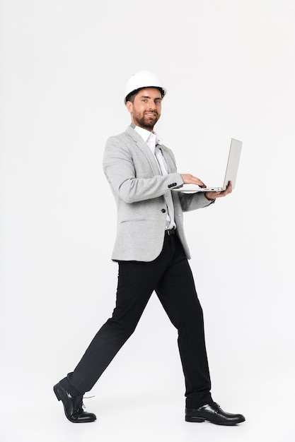 Full length of a confident bearded man builder wearing suit and hardhat standing isolated over white wall, using laptop computer