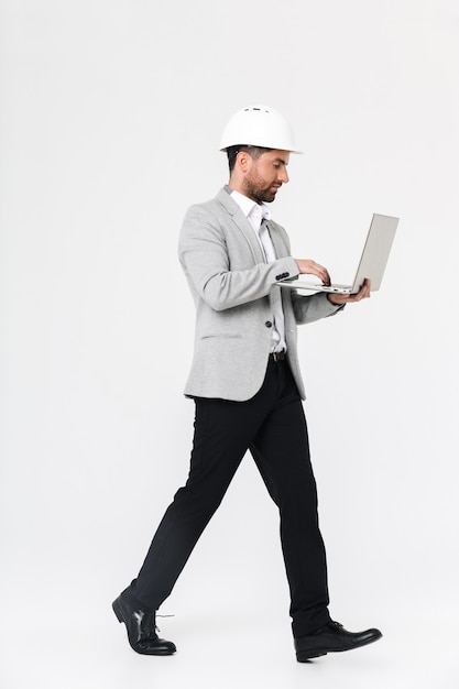 Full length of a confident bearded man builder wearing suit and hardhat standing isolated over white wall, using laptop computer