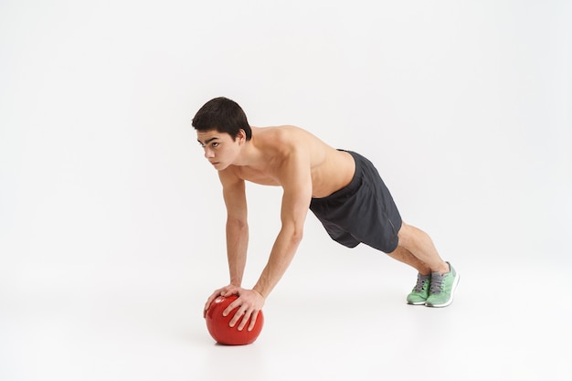 Молодой спортсмен без рубашки в полный рост делает упражнения с тяжелым мячом на белом