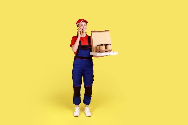 Full length of courier woman holds coffee and boxes with pizza, offering drinks and food delivery, talking via mobile phone with clients. Indoor studio shot isolated on yellow background