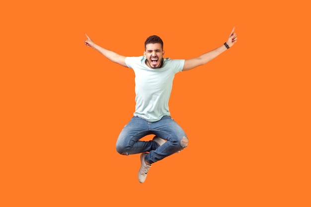 Full length of crazy overjoyed brunette man in white outfit jumping in air with raised hands, screaming loud for joy, feeling energetic and lively. indoor studio shot isolated on orange background