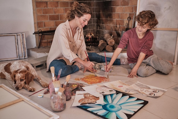 Full length of curly haired boy and female artist sitting on floor and looking down while coloring sheet of paper with paintbrushes at home near Spaniel dog