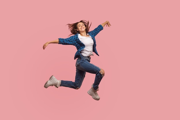 Full length of delighted happy stylish girl in checkered shirt and ripped jeans dancing in air celebrating success feeling free and inspired in flight indoor studio shot isolated on pink background