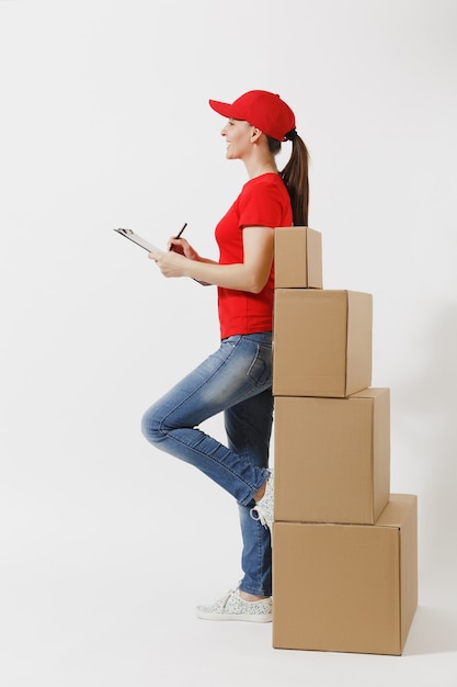 Full length of delivery woman in red cap, t-shirt isolated on white background. Female courier holding clipboard with papers document, blank empty sheet on empty cardboard boxes. Receiving package.