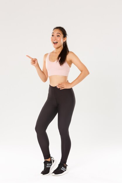 Full length of excited asian female athlete in sportsbra and leggings pointing and looking upper left corner thrilled.