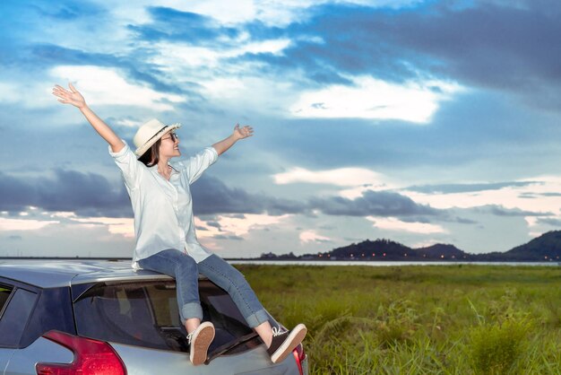 Full length f cheerful woman with arms raised sitting on car against sky