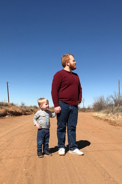 Photo full length of father and son standing on dirt road against clear blue sky