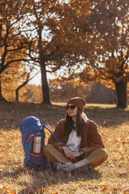 Full length of female traveler with backpack resting on meadow and enjoying autumn nature