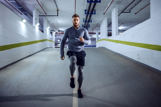 Full length of fit muscular handsome Caucasian sportsman in active wear running in underground garage at night. Urban life concept.