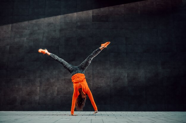 Full length of fit sporty caucasian woman in sportswear doing cartwheel exercise in front of black wall.