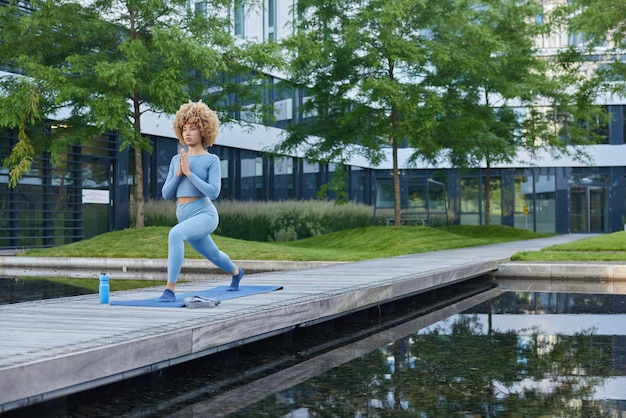 Full length of flexible woman stands on fitness mat practices
yoga wears sportswear breathes fresh air near lake in urban park
leads healthy lifestyle enjoys hatha practice for enlightenment
harmony