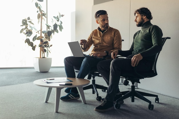 Полная длина сосредоточенных коллег-мужчин, использующих портативный компьютер в современном офисном пустом пространстве