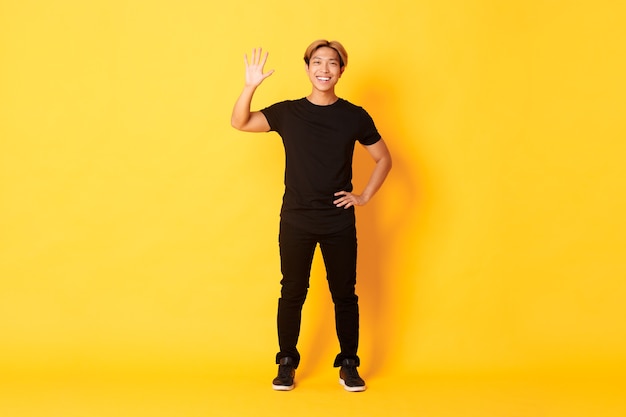 Full-length of friendly handsome asian man waving hand in hello, smiling and saying hi over yellow wall.