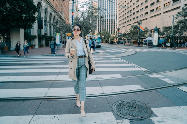 Full length front view beautiful asian korean woman crossing on
zebra crosswalk and holding take away coffee in hand. smiling young
office lady relax walking in city street. gay flag hanging
usa.