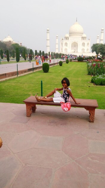 Photo full length of girl sitting on bench against taj mahal