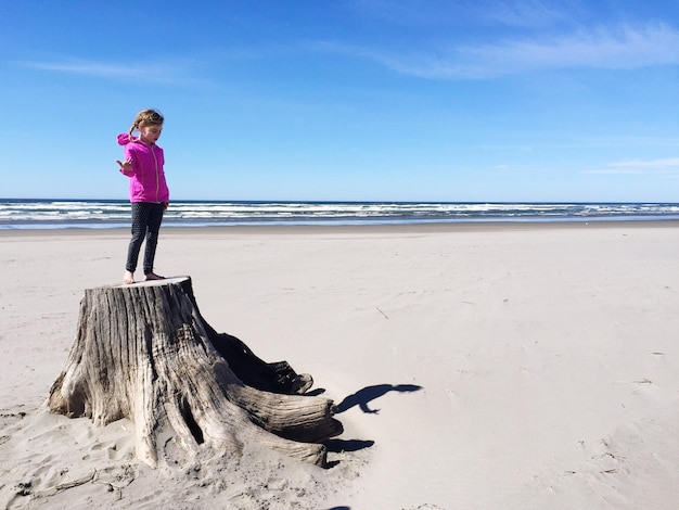 Foto lunghezza completa di una ragazza in piedi sul tronco di un albero sulla spiaggia contro il cielo