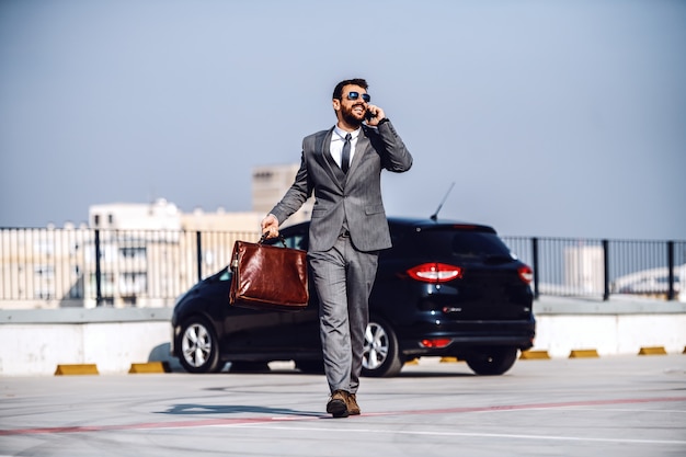 Photo full length of handsome caucasian bearded businessman in suit and with sunglasses walking on parking lot, talking on the phone and holding briefcase.