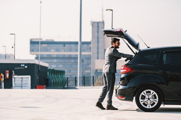 Full length of handsome caucasian bearded rich businessman in suit opening trunk on his car and putting his briefcase in it.