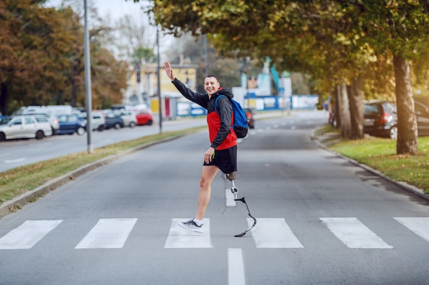 スポーツウェアのハンサムな白人の障害者スポーツマンの全長。義足とバックパックが通りを横切り、友達に手を振っています。