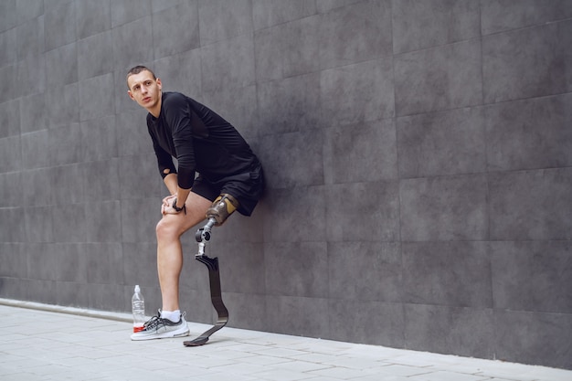 В полный рост красивый кавказский спортсмен с искусственной ногой опирается на стену и отдыхает от бега. Рядом с ним бутылка с прохладительными напитками.