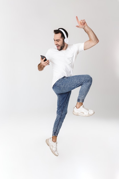 Per tutta la lunghezza di un bell'uomo allegro che indossa una t-shirt bianca in piedi isolato su un muro bianco, ascoltando musica con le cuffie, usando il telefono cellulare