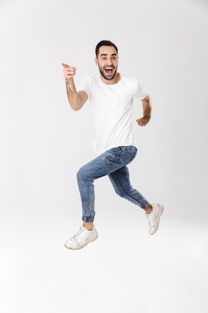 Full length of a handsome cheerful man wearing blank t-shirt standing isolated over white wall, pointing finger, jumping