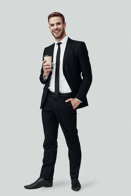 Full length of handsome young man in formalwear looking at camera and smiling while standing against grey background