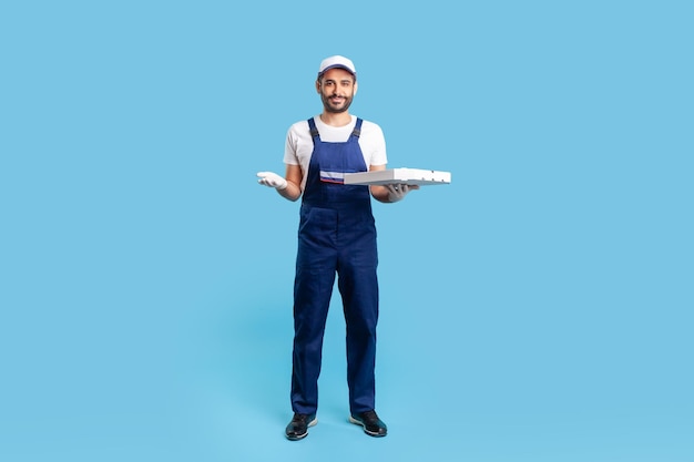 Full length happy courier in workwear and protective gloves carrying pizza box holding empty space on palm for advertise image Fast food delivery service studio shot isolated on blue background