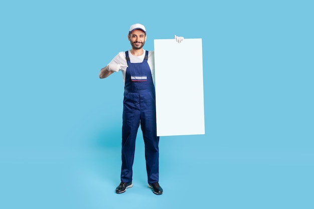 Full length of happy labourer man in uniform and protective gloves pointing at vertical poster template mock up blank space for advertising message recommendations and tips studio shot isolated