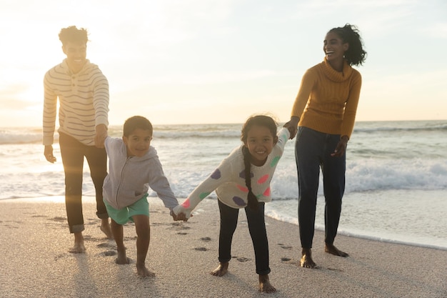 Full length of happy multiracial children holding hands pulling parents at beach against sky