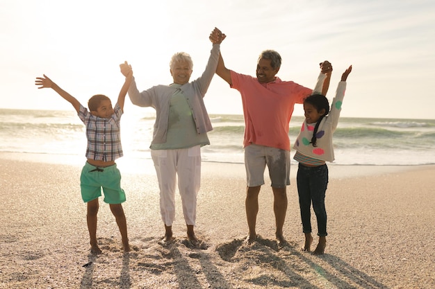 Full length of happy multiracial grandparents and grandchildren holding hands at beach against sky