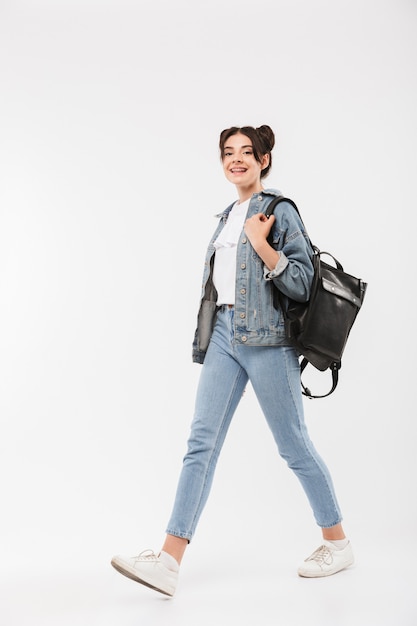 Full length happy student girl with double buns hairstyle wearing jeans clothing and backpack walking with smile, isolated on white