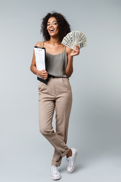 Photo full length of a happy young african woman casually dressed standing isolated, holding a notepad, showing money banknotes