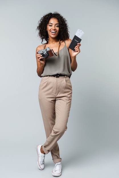 Full length of a happy young african woman casually dressed standing isolated, holding photo camera, showing passport with flight tickets